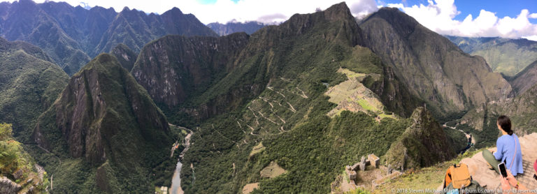 Visitor at a Huayna Picchu overlook with Machu Picchu far below among steep jagged Andean mountains.
