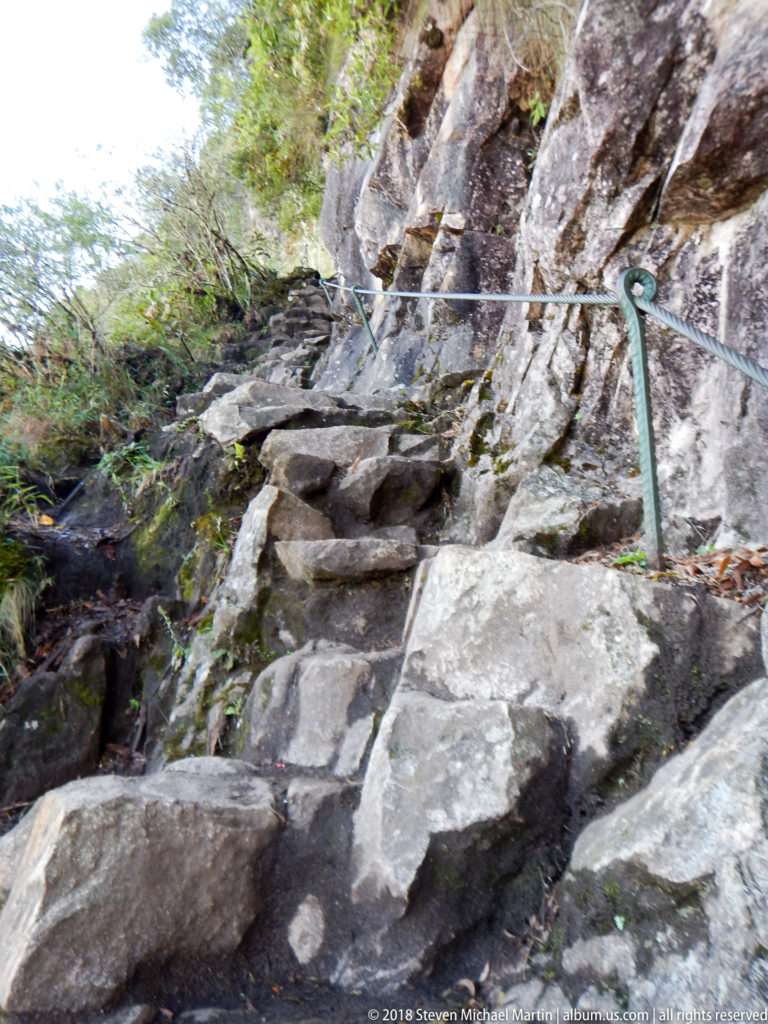 Steep uneven stone steps with a metal handrail climb between exposed rock and vegetation on Huayna Picchu.