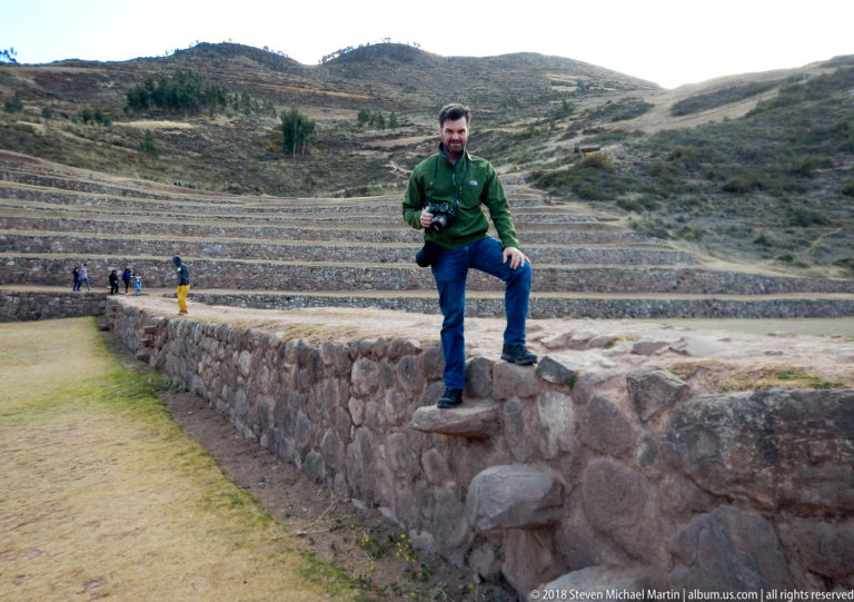 Man with a camera standing on projecting stone steps in a Moray terrace wall, with curved terraces behind.