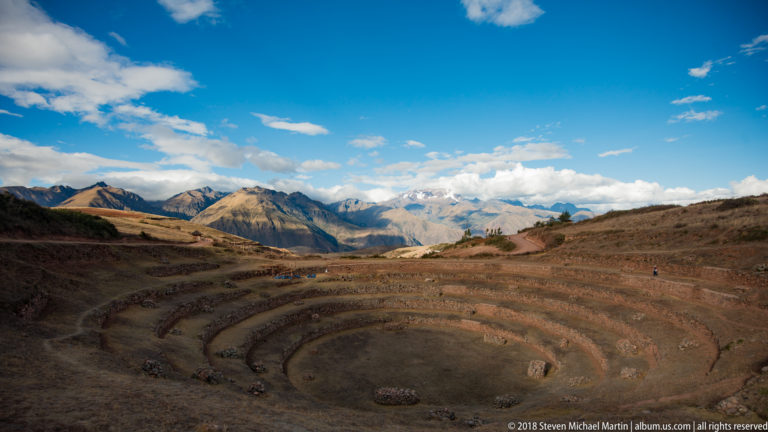 Moray's circular terraces below a wide blue sky and distant rugged Andean mountains.