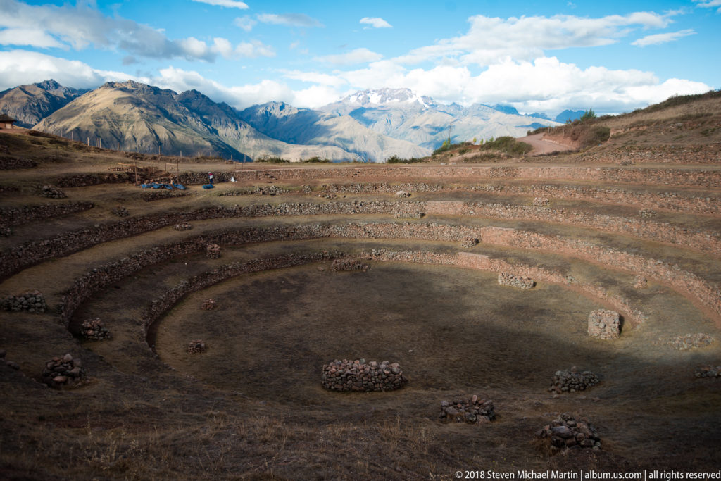 Curved Moray terraces and stone piles in the foreground with layered Andean mountains extending across the distant horizon.