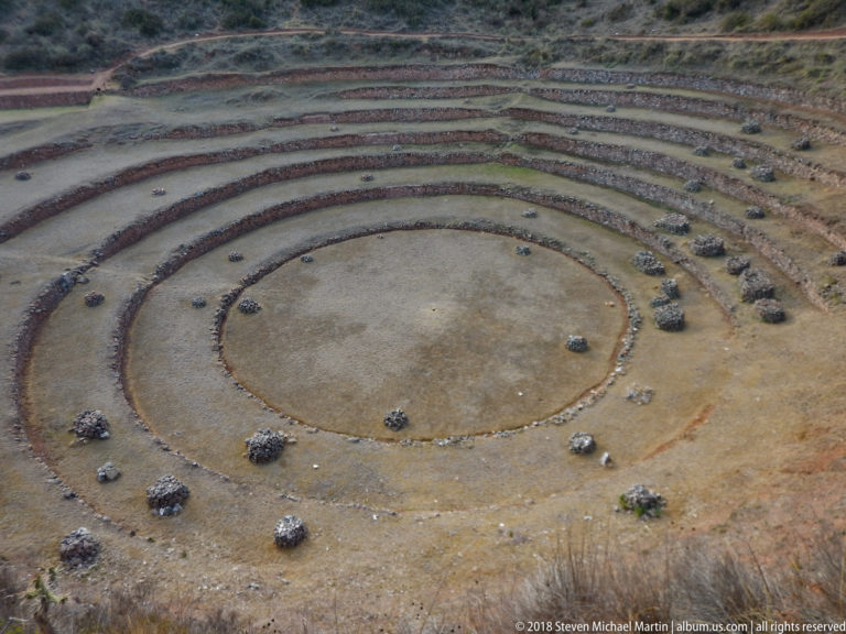 Elevated view into Moray's circular center, surrounded by concentric low stone walls and scattered piles of stones.