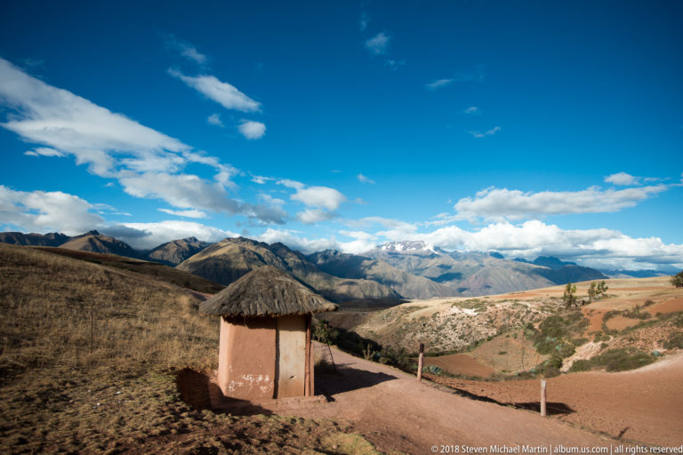 Small thatched shelter beside a reddish path overlooking dry Andean hills, patchwork fields, and distant clouded mountains.