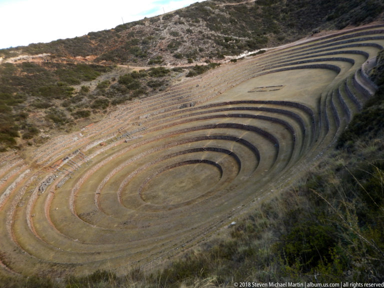 Large concentric terraced bowl at Moray viewed obliquely, with alternating sunlit and shaded rings below scrub-covered slopes.