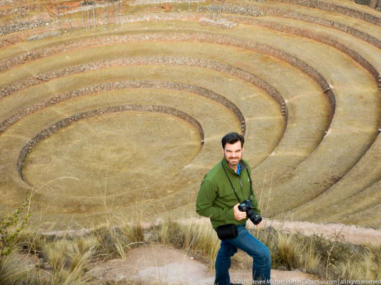 Man in a green jacket holding a camera at the rim above Moray's broad concentric terrace rings.