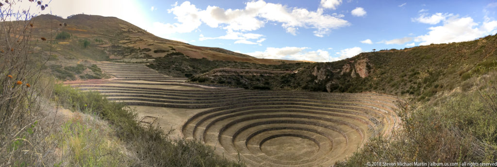 Moray's concentric terraces seen through tall foreground grasses, with hillside terraces, rocky slopes, and blue sky beyond.