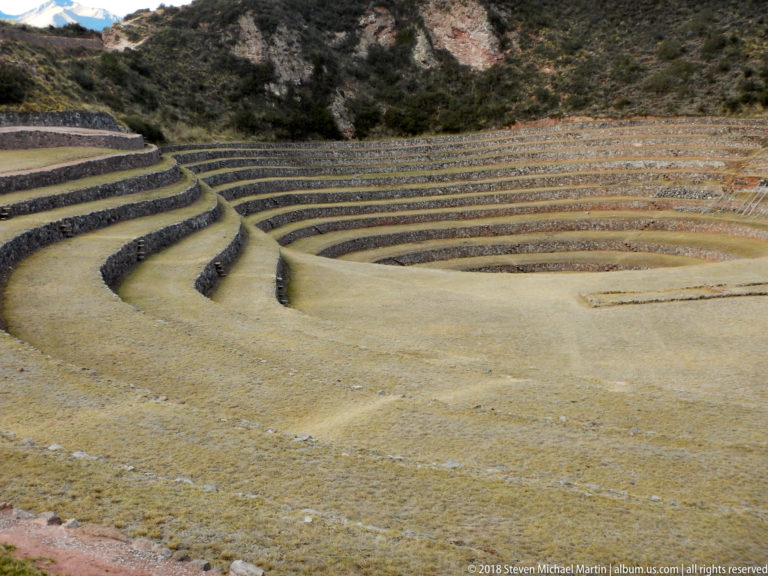 Broad central floor at Moray bordered by curving stone-edged terraces rising toward a rocky, shrub-covered slope.