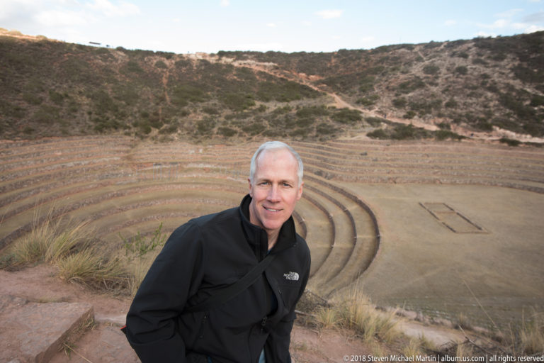 Man in a black jacket standing at an overlook above Moray's concentric terraces and scrub-covered hillside.