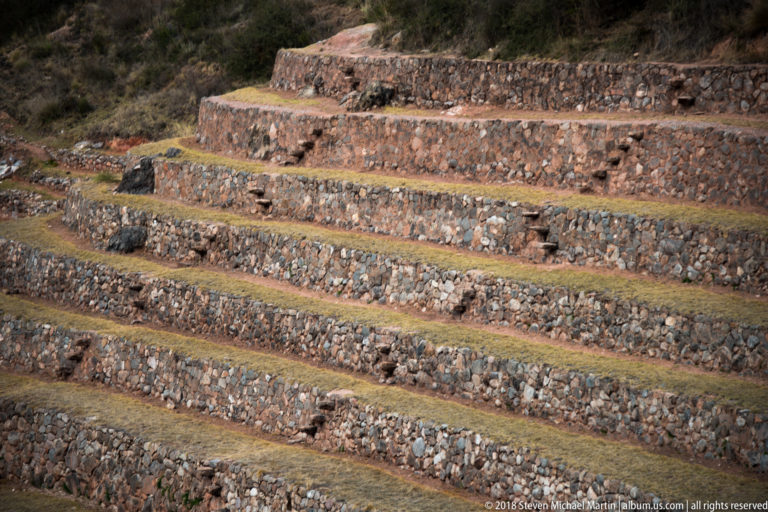 Sunlit stacked stone retaining walls and dry grass terrace benches at Moray, with projecting stones between levels.