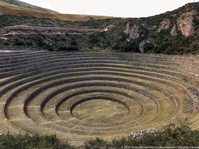 Tightly framed concentric terraces at Moray descending toward a circular center beneath a rocky shrub-covered slope.