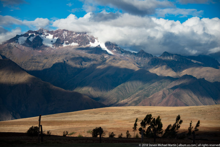 Snowcapped Andean peaks beneath clouds beyond layered dark ridges, with a pale field and scattered trees in front.