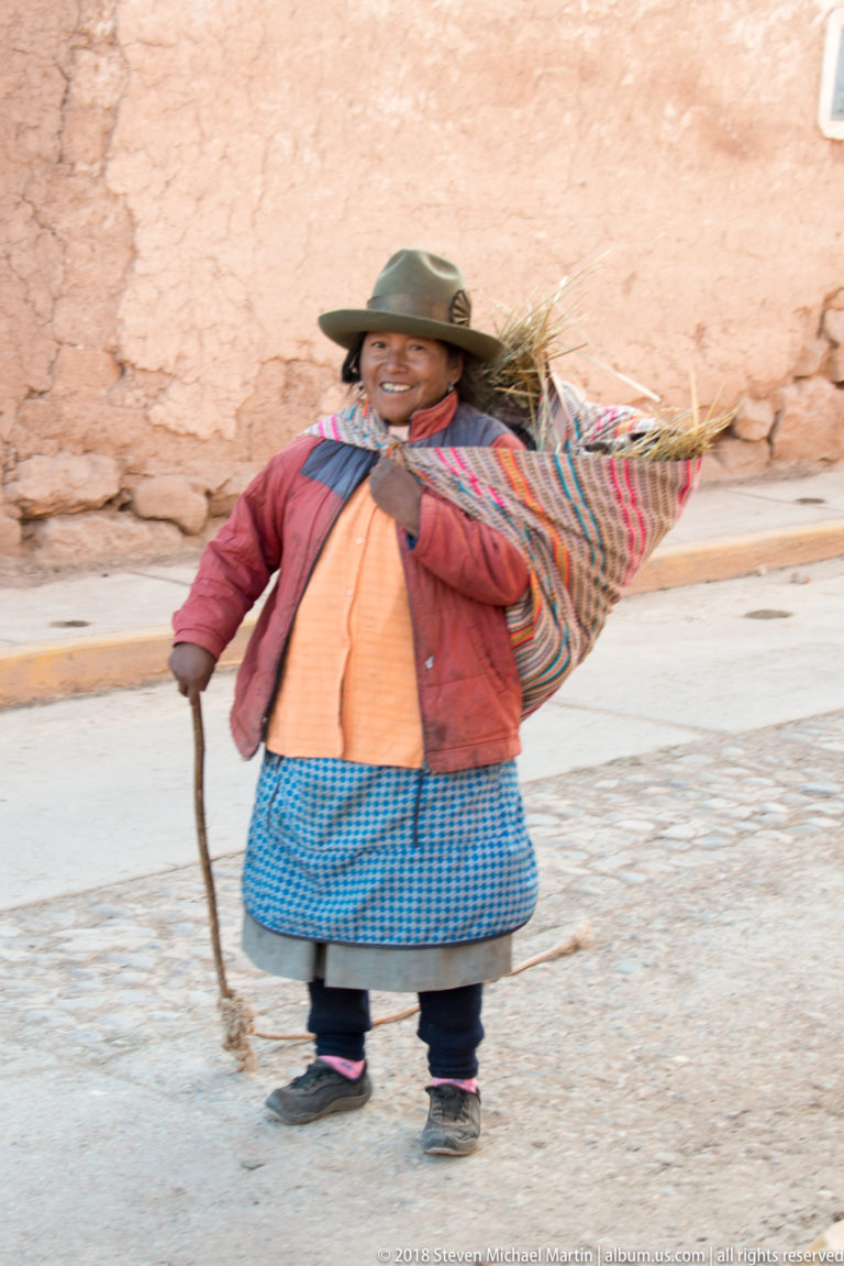 Full-length view of a woman in layered clothing carrying dry grass and holding a stick beside an earthen wall.