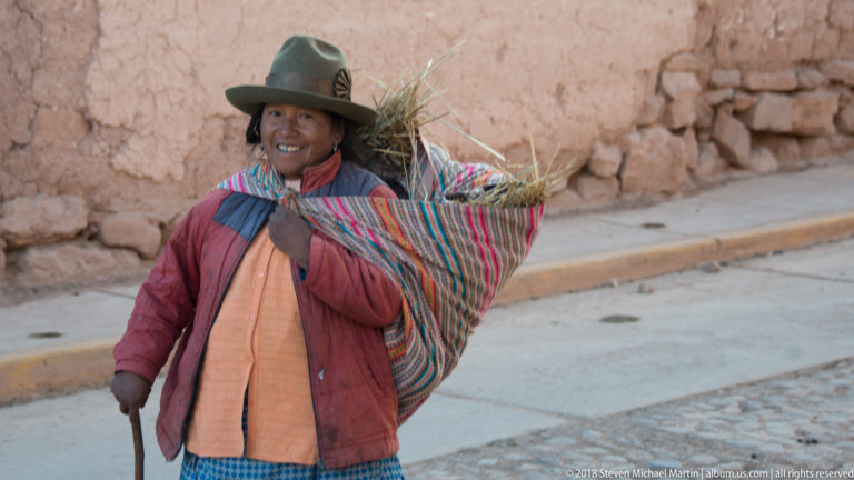 Woman beside an earthen wall carrying dry grass in a striped woven cloth and holding a long walking stick.