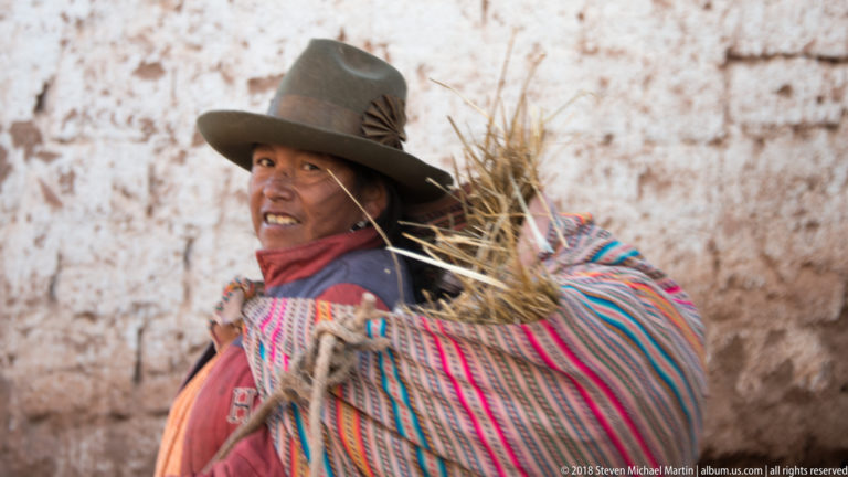 Smiling woman in a broad hat carrying dry grass in a brightly striped woven cloth against a pale wall.