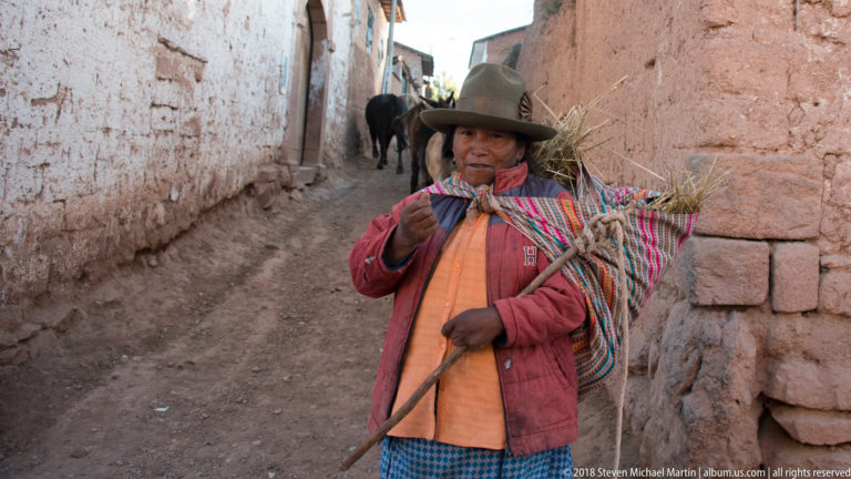 Woman in a brimmed hat carrying dry grass in a colorful woven cloth along a narrow earthen-walled lane.