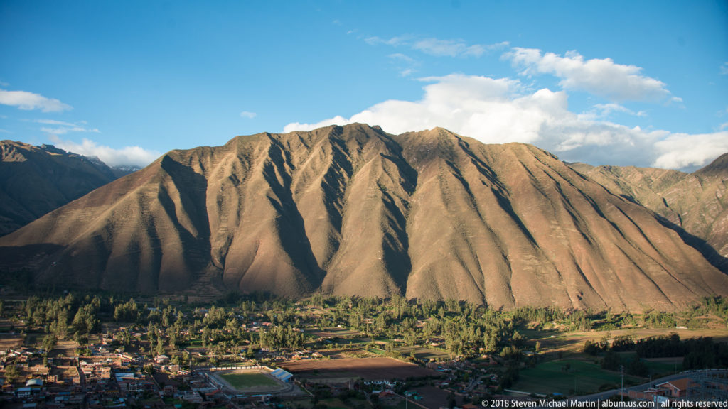Sunlit, deeply furrowed Andean ridge rising above green fields, trees, and buildings beneath a blue sky.