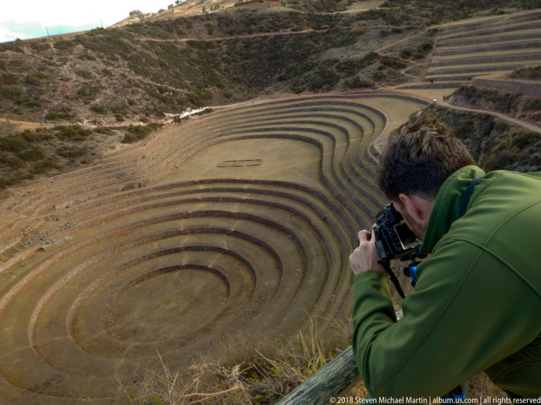Man in a green jacket photographing Moray's concentric terraces from a high overlook.
