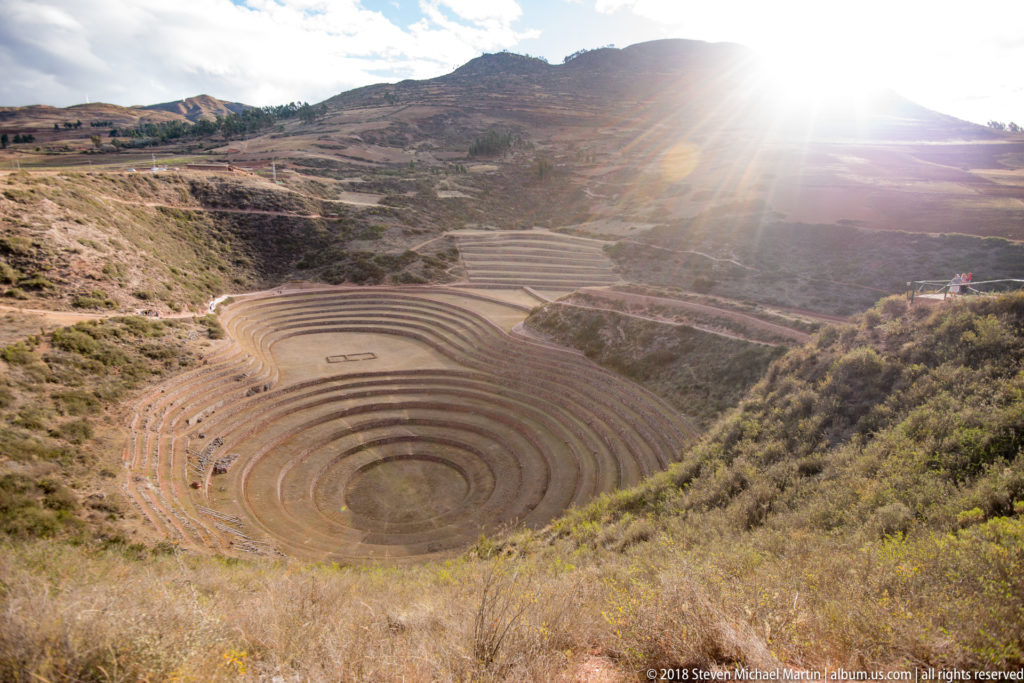 Backlit high view of Moray's circular terraces and far hillside levels, with bright sun flare above the rim.