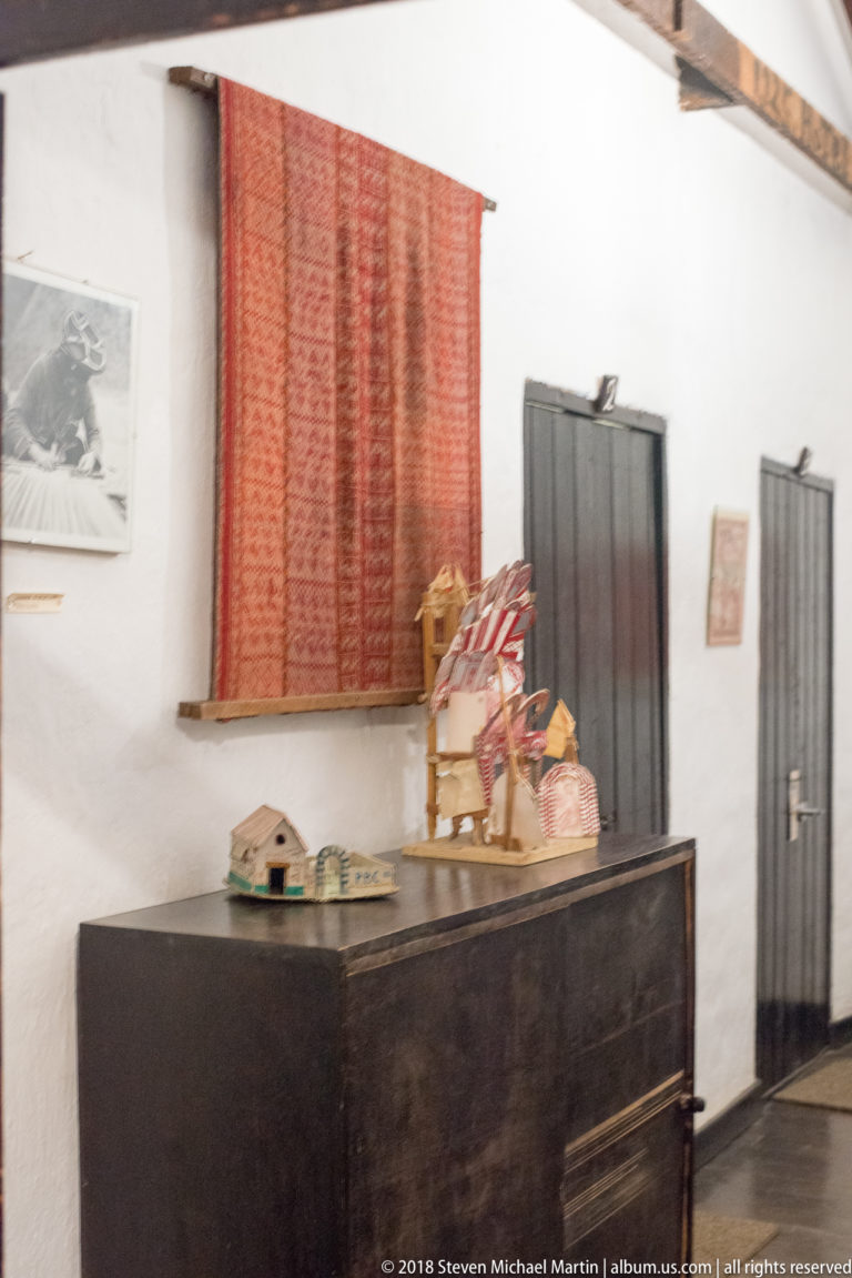 White-walled rustic interior with dark wooden chest, red woven textile, small craft objects, and black plank doors.
