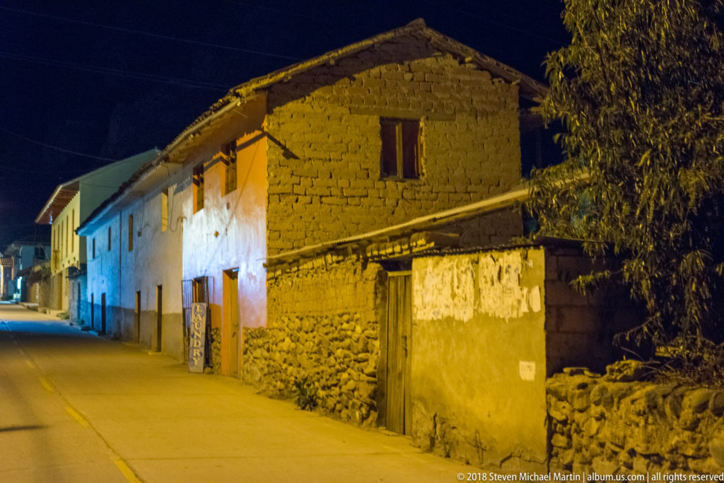 Quiet nighttime street lined with adobe, plaster, and stone buildings under warm yellow and blue streetlights.