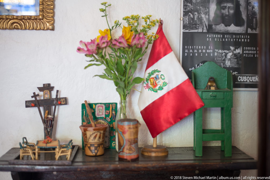Peruvian flag beside flowers, small craft objects, painted vessels, and an Ollantaytambo poster on a tabletop.