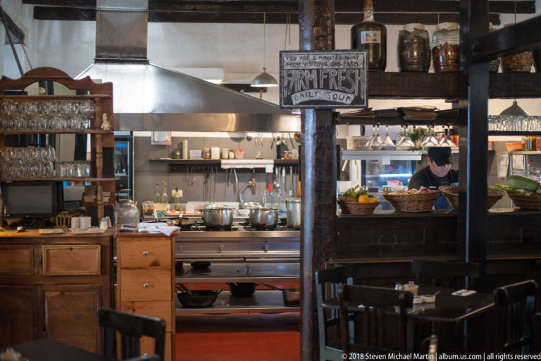 Open restaurant kitchen with stainless-steel equipment, glass shelves, wooden cabinetry, and a cook at work.