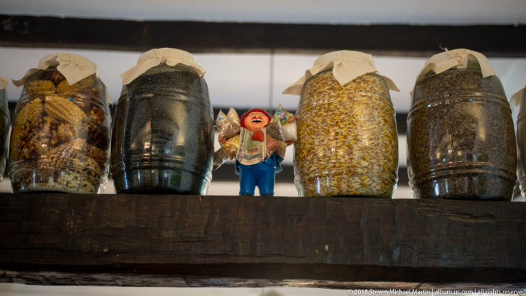 Glass jars filled with dried corn, grains, or seeds on a wooden shelf beside a small colorful figurine.