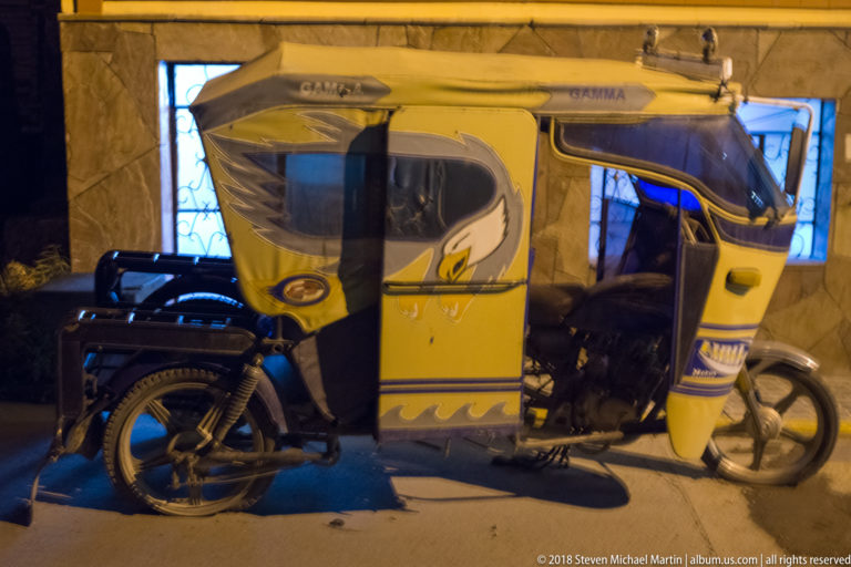Side view of a yellow three-wheeled mototaxi with covered passenger cabin parked at night.
