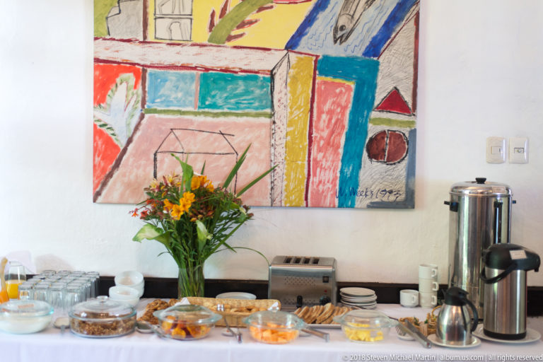 Breakfast buffet with juice, cereals, dishes, flowers, and hot-water urn beneath a colorful abstract painting.
