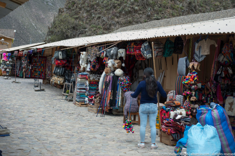 Row of market stalls displaying textiles, bags, plush alpacas, and colorful souvenirs along a cobbled lane.