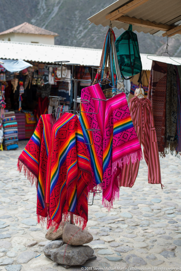 Bright red and magenta woven Andean textiles with stripes, geometric motifs, and fringe hanging at a market.