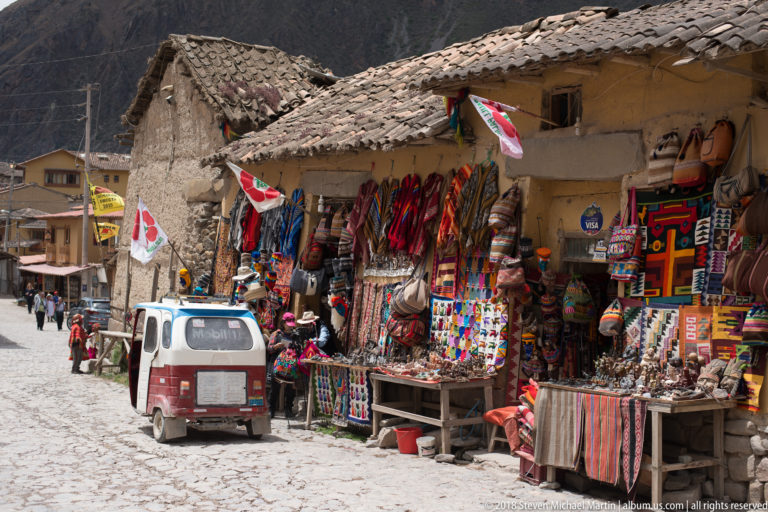 Cobbled street lined with colorful textile and handicraft shops, with a small vehicle passing through.