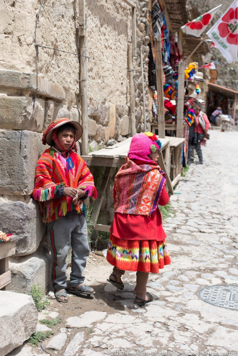 Two people in colorful Andean clothing beside a stone wall on a cobbled street lined with souvenir stalls.