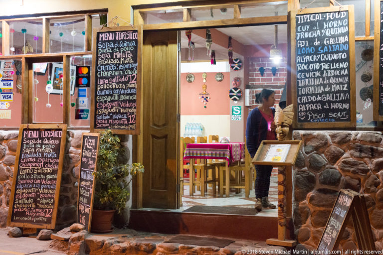 Stone-fronted restaurant entrance crowded with chalkboard menus advertising typical Peruvian food.