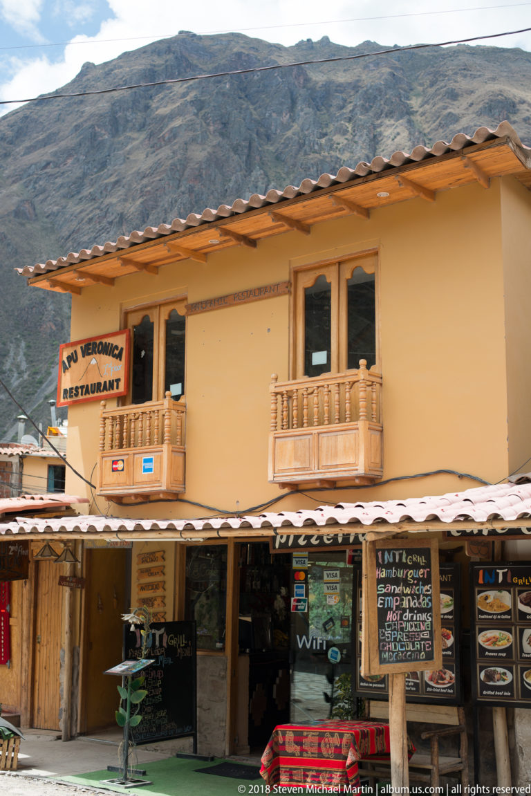 Yellow two-story restaurant with wooden balconies, menu boards, awning, and steep mountain slope behind.