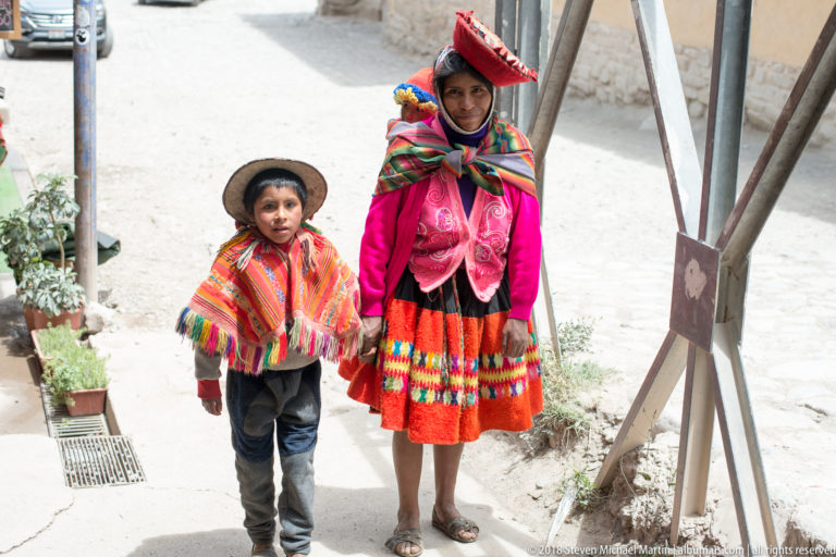 Woman and young child in colorful Andean clothing looking up toward the camera on a sunlit street.