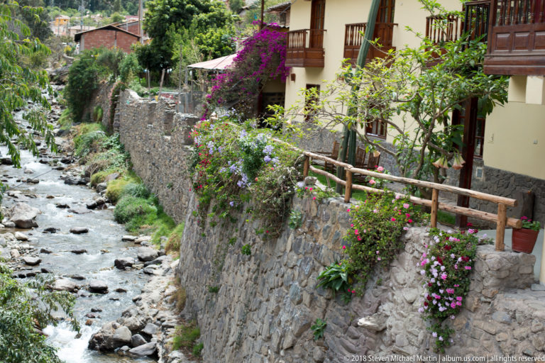 Close view of a rocky canal beside stone walls, flowering plants, and timber balconies.