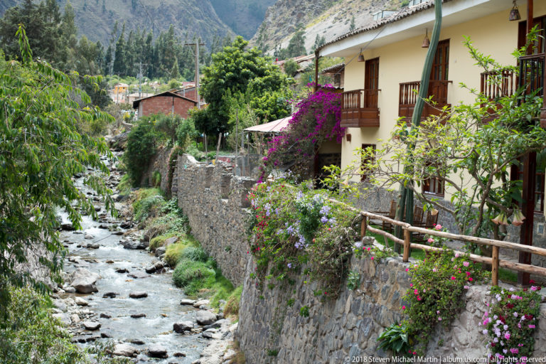 Stone-lined canal flowing beside flower-covered walls and houses with wooden balconies in Ollantaytambo.