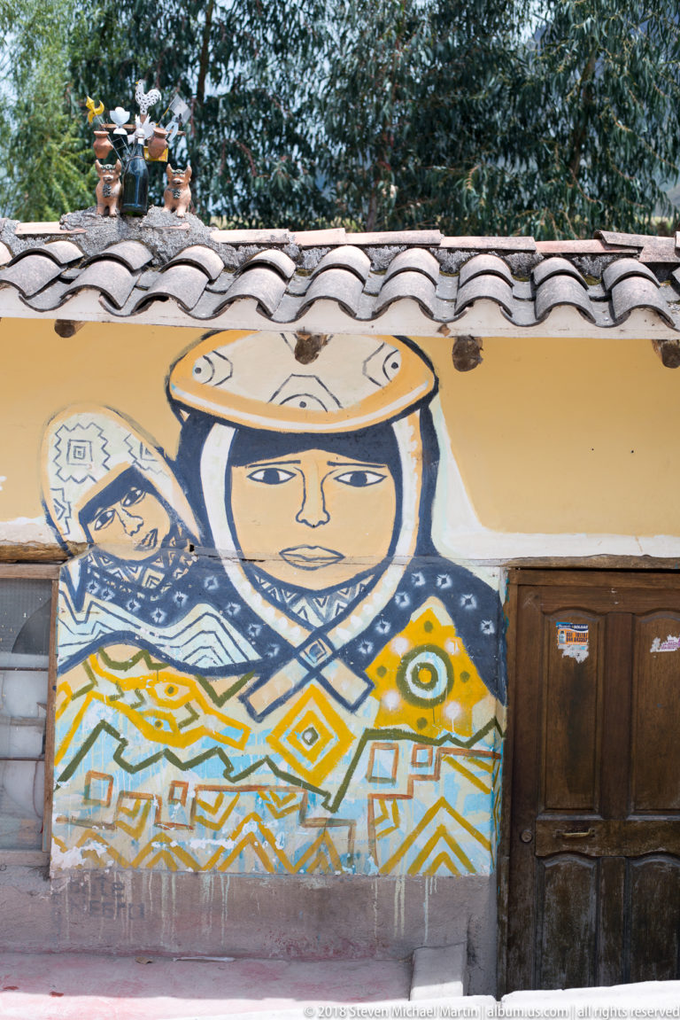 Painted wall mural with stylized faces and colorful geometric motifs beneath a tiled roof and beside a wooden door.