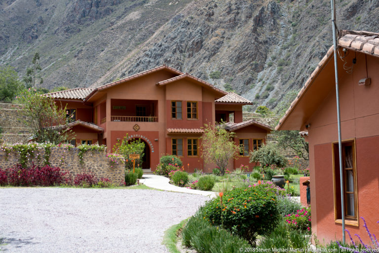 Terracotta lodge buildings with tiled roofs and gardens set below steep rocky Andean mountains.