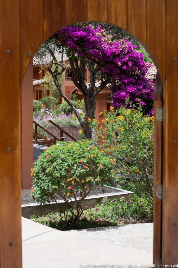 Arched wooden doorway framing a garden with purple bougainvillea, flowering shrubs, paths, and terracotta-colored buildings.