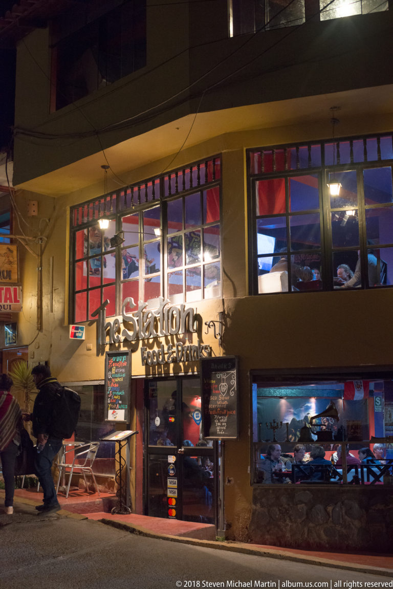 Night exterior of The Station restaurant with colorful upper windows, chalkboard menus, and diners visible inside.
