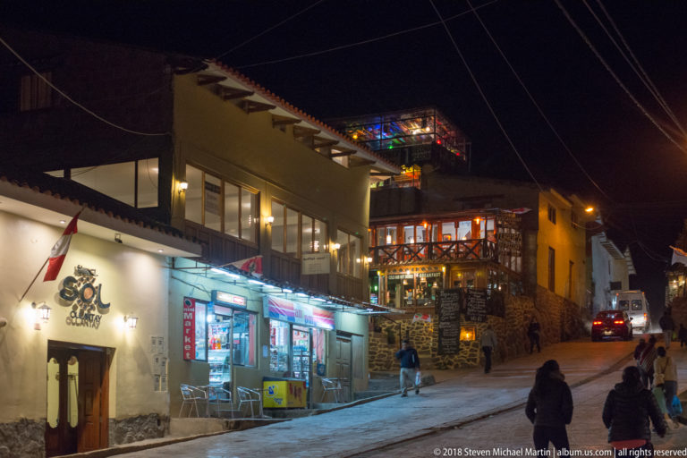 Uphill street at night lined with illuminated restaurants and shops, with pedestrians and overhead wires.