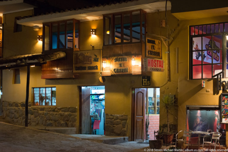 Yellow two-story Hostal Chaska façade at night with illuminated signs, open doors, and stone lower walls.