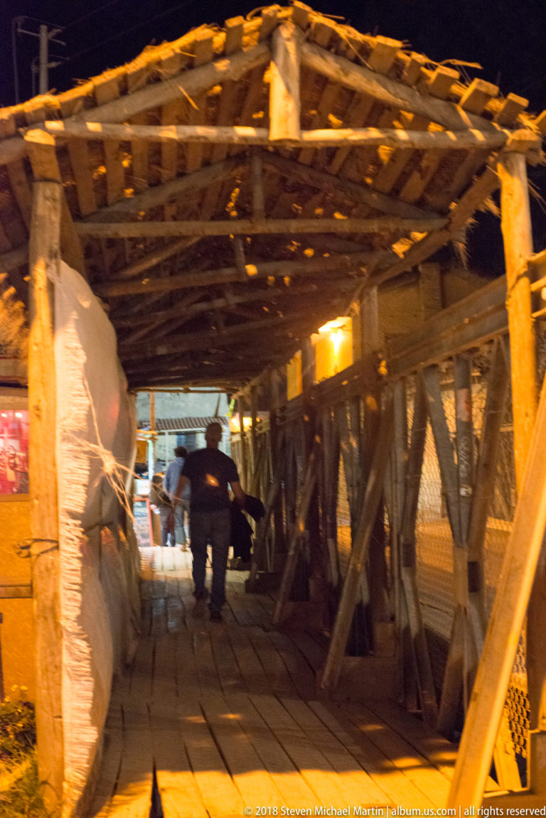 Rustic covered wooden passage with heavy posts, diagonal braces, and a person walking away at night.