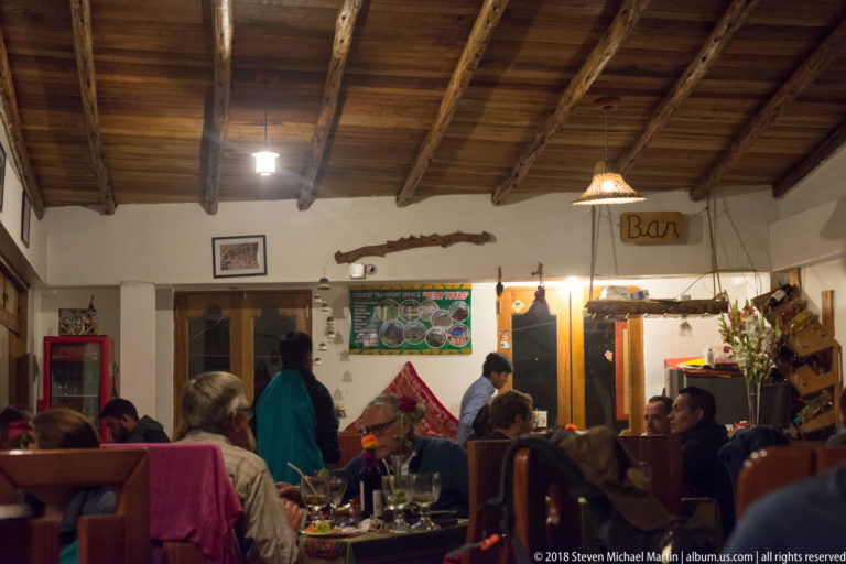 Busy restaurant dining room with exposed timber ceiling, pendant lights, diners, and a small bar area.