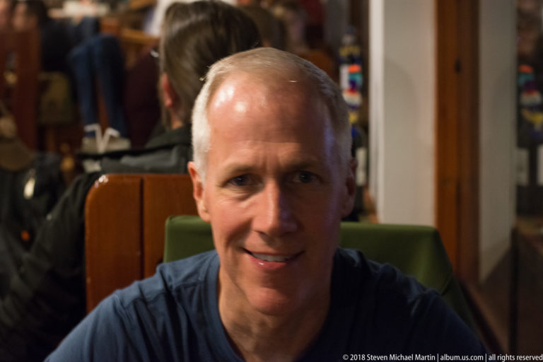 Adult man seated and smiling slightly in a restaurant, with other diners softly blurred behind.