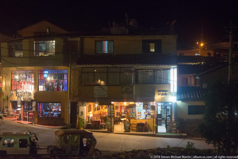 Night view across a street toward illuminated restaurants, shops, signs, and parked mototaxis.