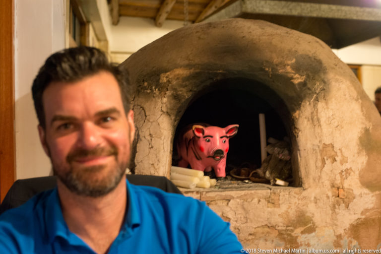 Close portrait of man beside a cracked domed clay oven containing a pink pig figure.