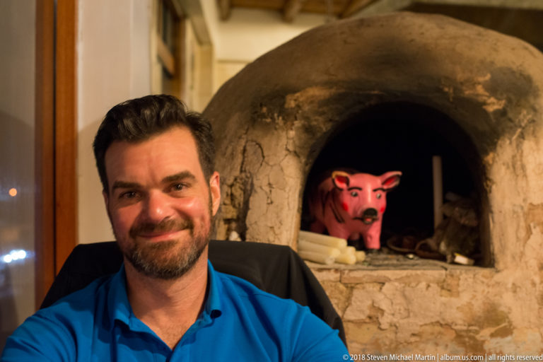 Man in a blue shirt seated beside a domed clay oven with a bright pink pig figure inside.